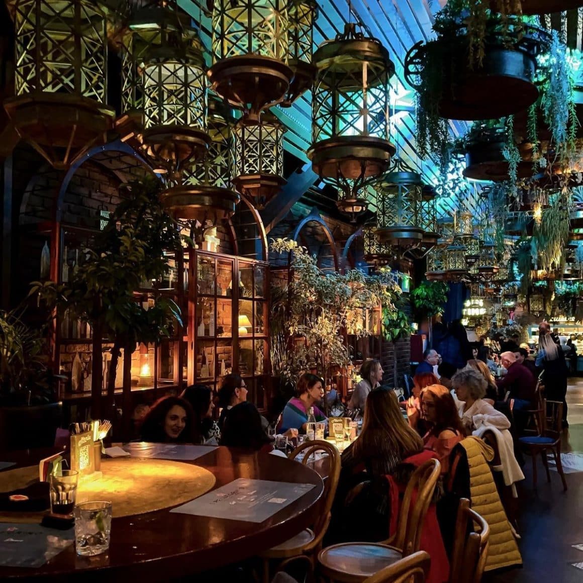 mai multi tineri socializează la mesele unui restaurant pentru grupuri mari din Bucuresti, cu multe plante curgătoare și multe candelabre asezate la diferite inălțimi
