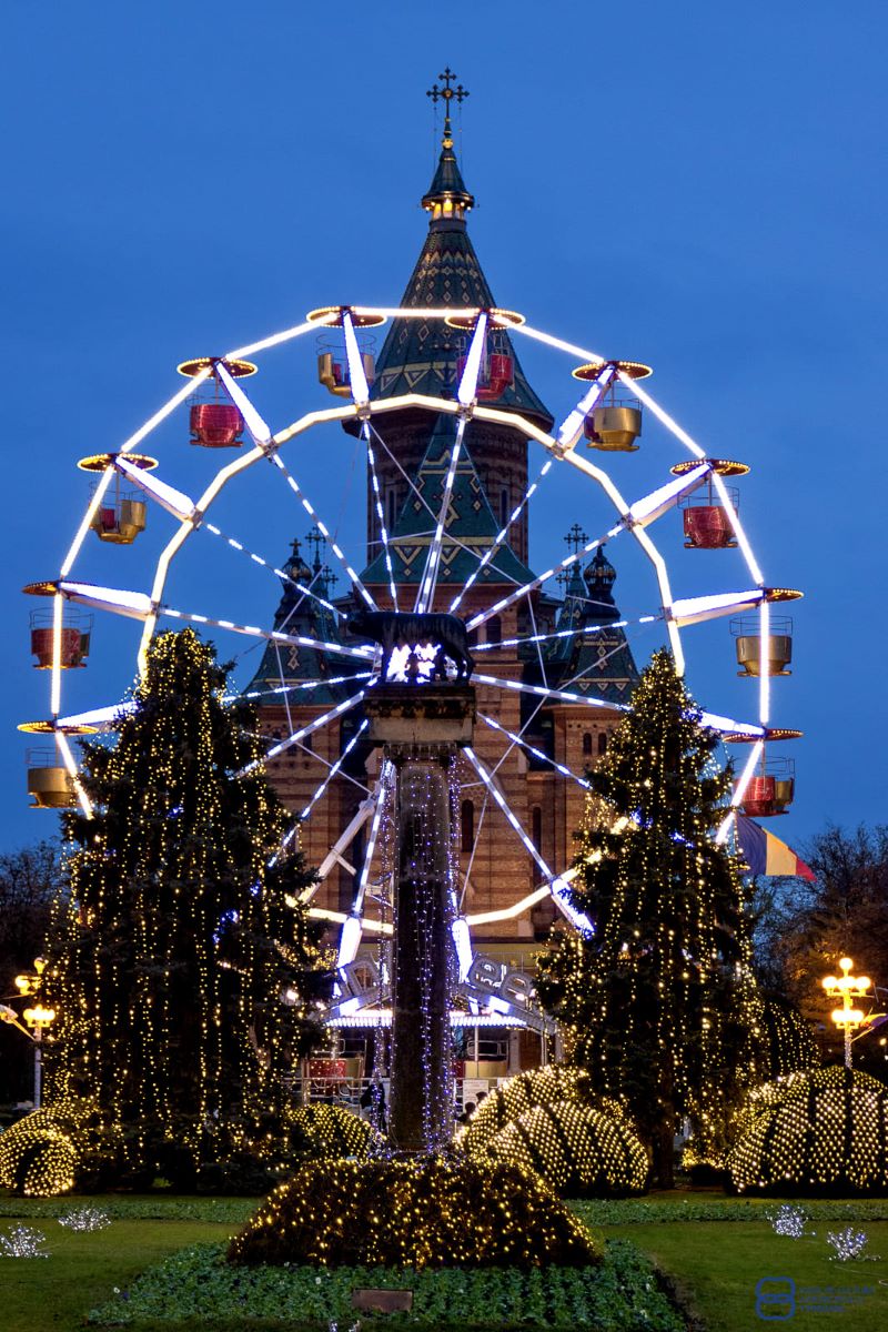 o fotografie cu rata din targul de crăciun din timișoara, cu doi brazi împodobiți lângăși o roată mare, luminată