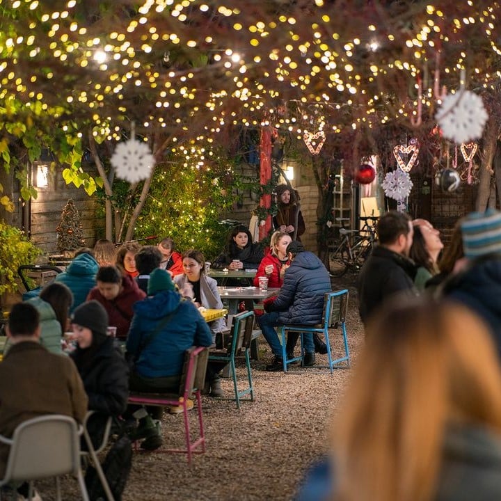 mai mulți tineri stau la mesele din curtea jai bistrot din bucuresti, iar deasupra meselor sunt atârnate ghirlande luminoase