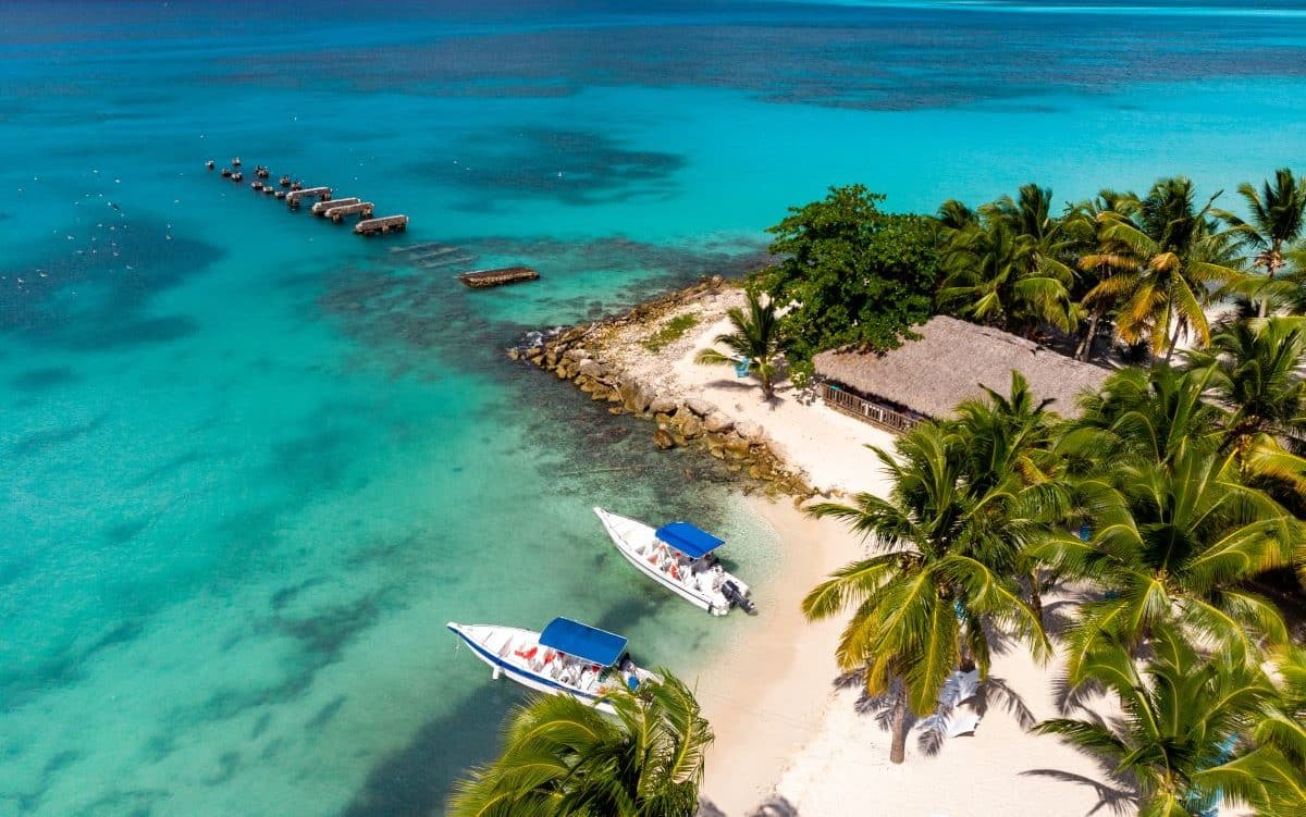 un peisaj exotic cu o terasă acoperită, două ambarcațiuni, mai multi palmieri, și oceanul în fundal. Punta Cana. Republica Dominicană. destinații exotice de Revelion