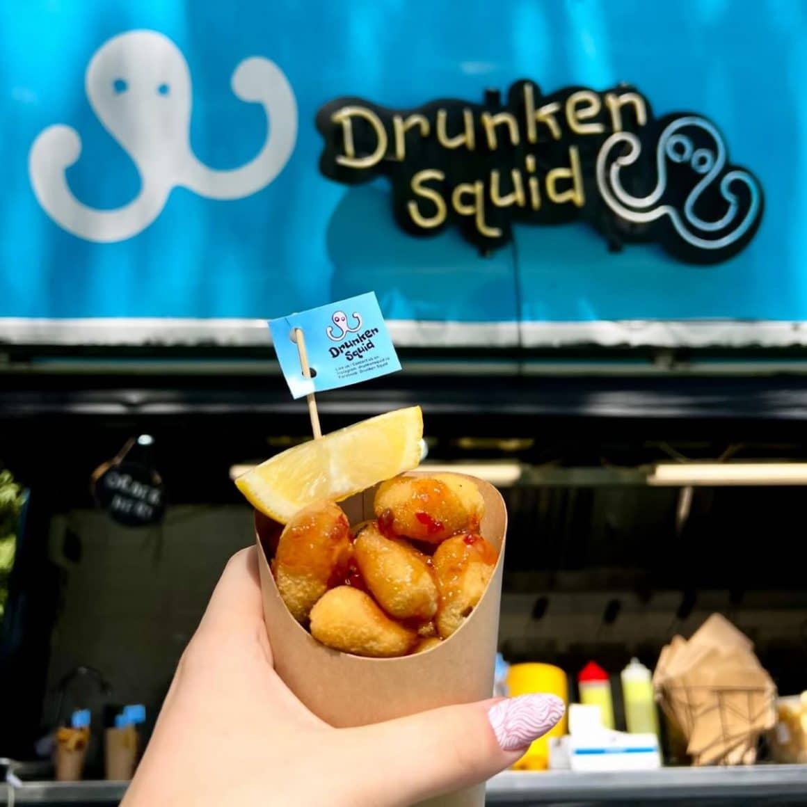 o femeie târnără ține în mână un recipient cu măsline pane în fața unui food truck la un festival