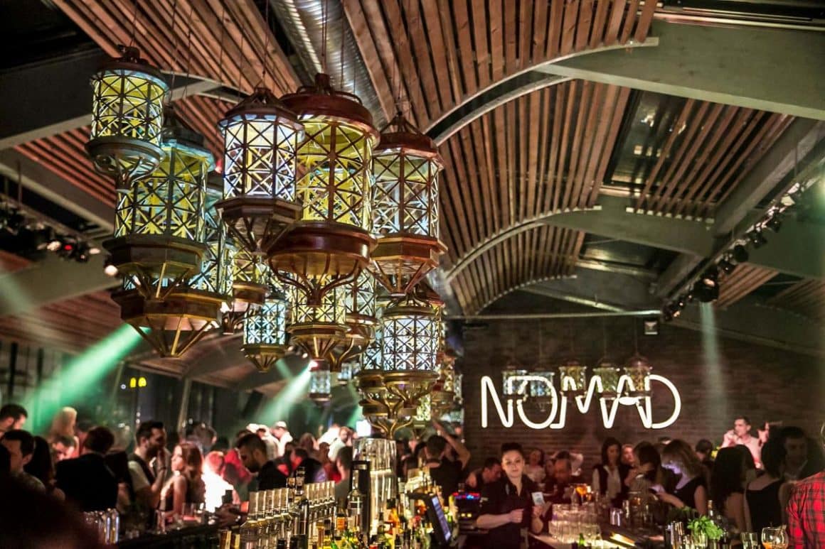 mai multi tineri socializează la mesele unui restaurant pentru grupuri mari din Bucuresti, cu tavan din riflaje din lemn și multe candelabre asezate la diferite inălțimi