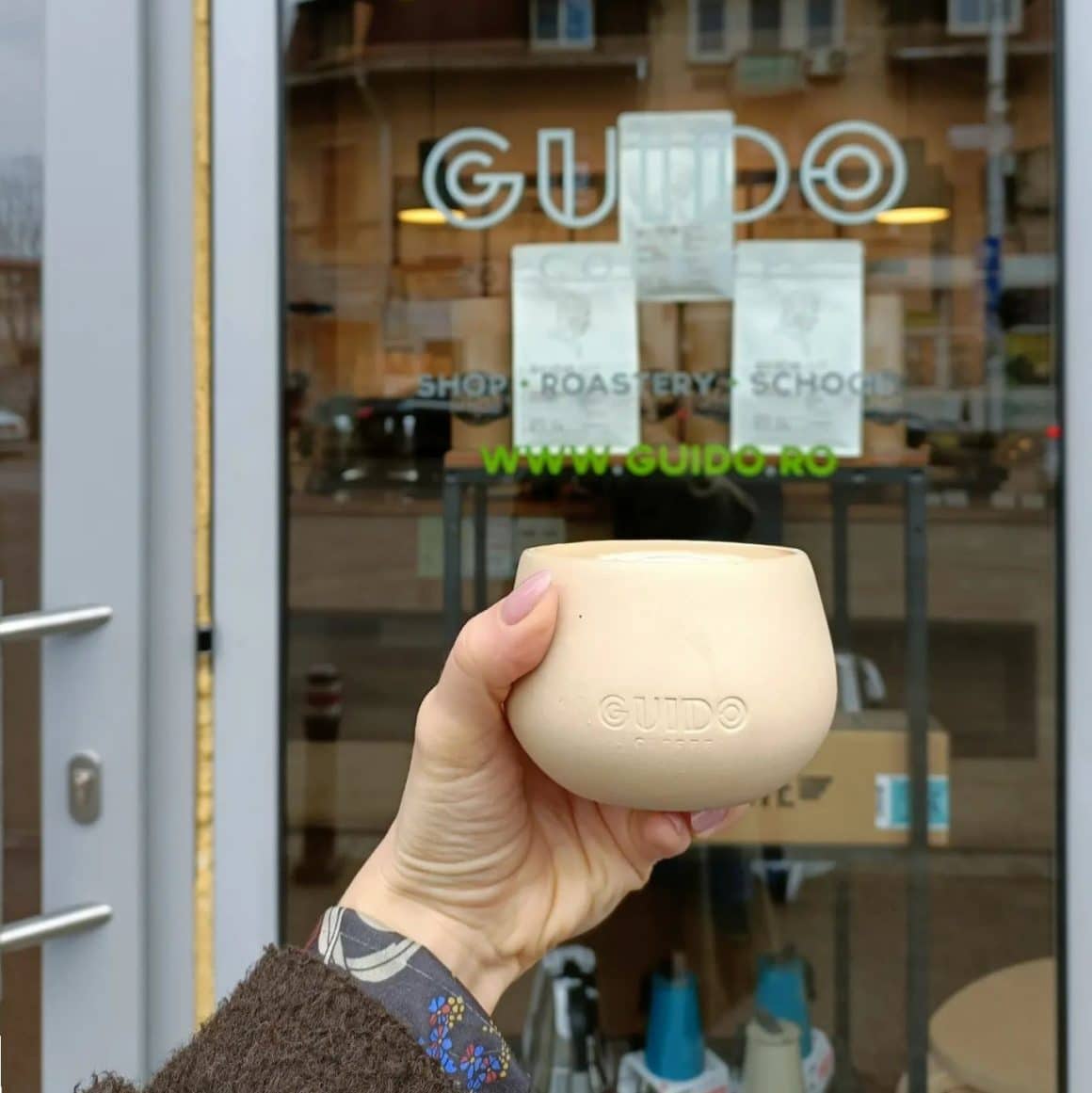 o femeie tine in mână o ceașcă de cafea, în fața intrării in cafenea