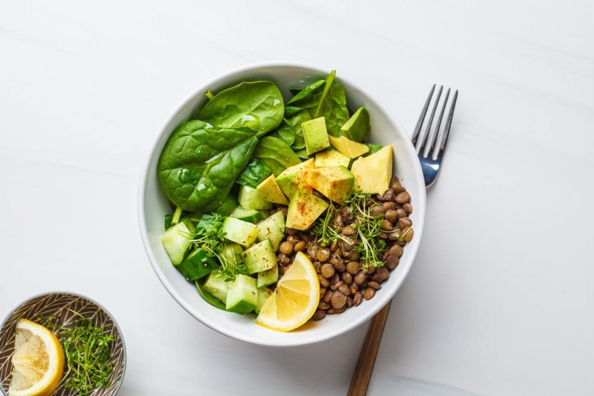 un bol cu mai multe ingrediente folosite la prepararea unei salate calde salate calde, așezat pe o față de masă albă, alături de un alt bol cu o felie de lămâie