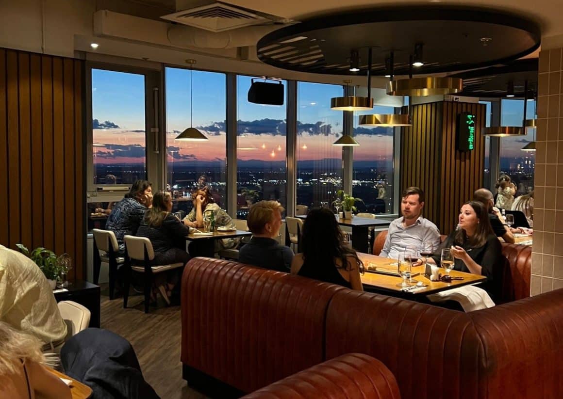 imagin din restaurantul la înălțime NOR din București, fotografiat la apus, cu mese si canapele si geamuri mari prin care se vedere orasul la venirea serii