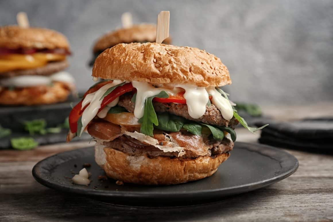 3 burgeri pe farfurii negre, cu prosciutto, branza, rosii si salata. Toppinguri pentru burgeri.