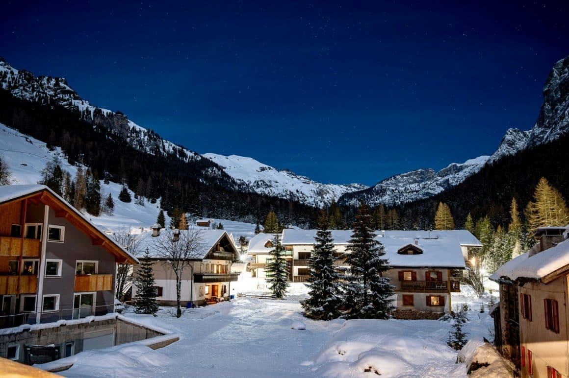 frup de mai multe pensiuni fotografiate iarna, acoperita de zapada, seara, iluminate, cu muntii in fundal.
