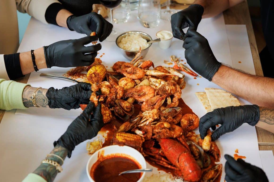 close up cu o masa pe care sunt rasturnate mai multe fructe de mare in sos cajun, din care oamenii mancanca cu mâna