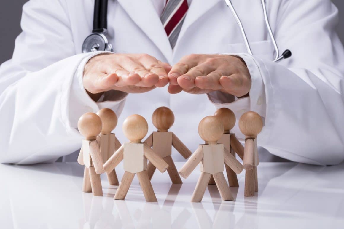 imagine concept pentru asigurari medicale: un doctor tine palmele deasupra unor omuleti din lemn