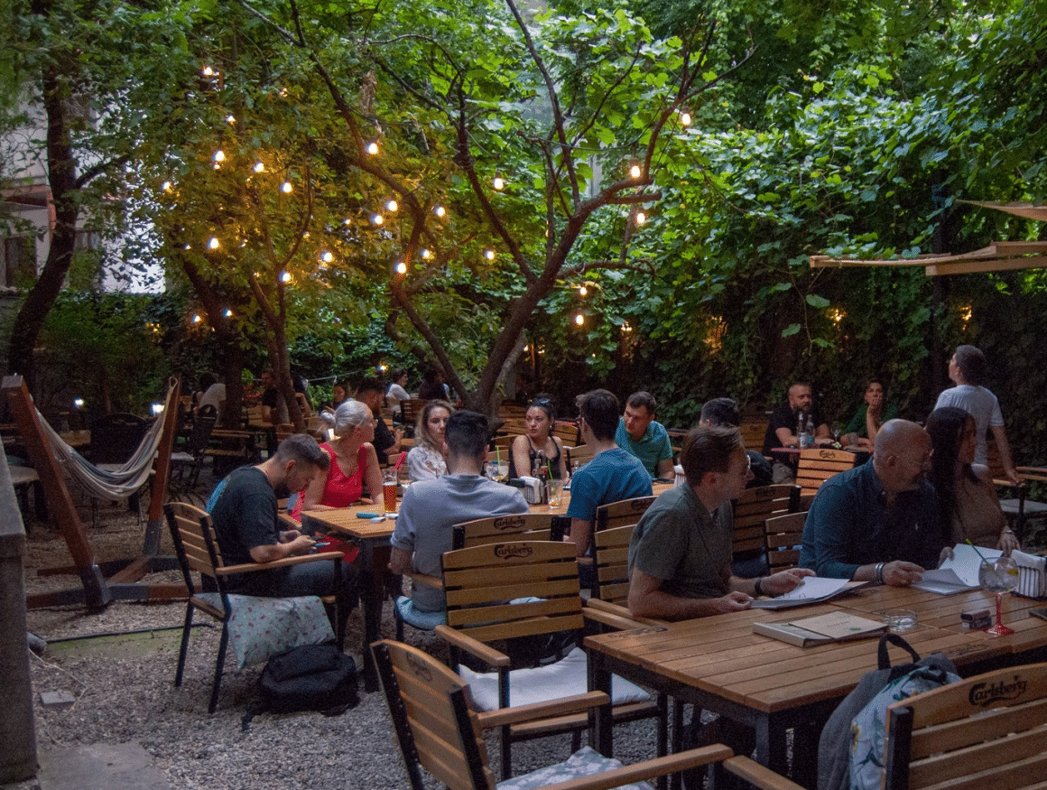 terasa Lente Arcului din București, cu mesele pline, de o parte si alta a unei alei, cu ziduri portocalii luminate cu aplice si deasupra multa verdeata de la copaci