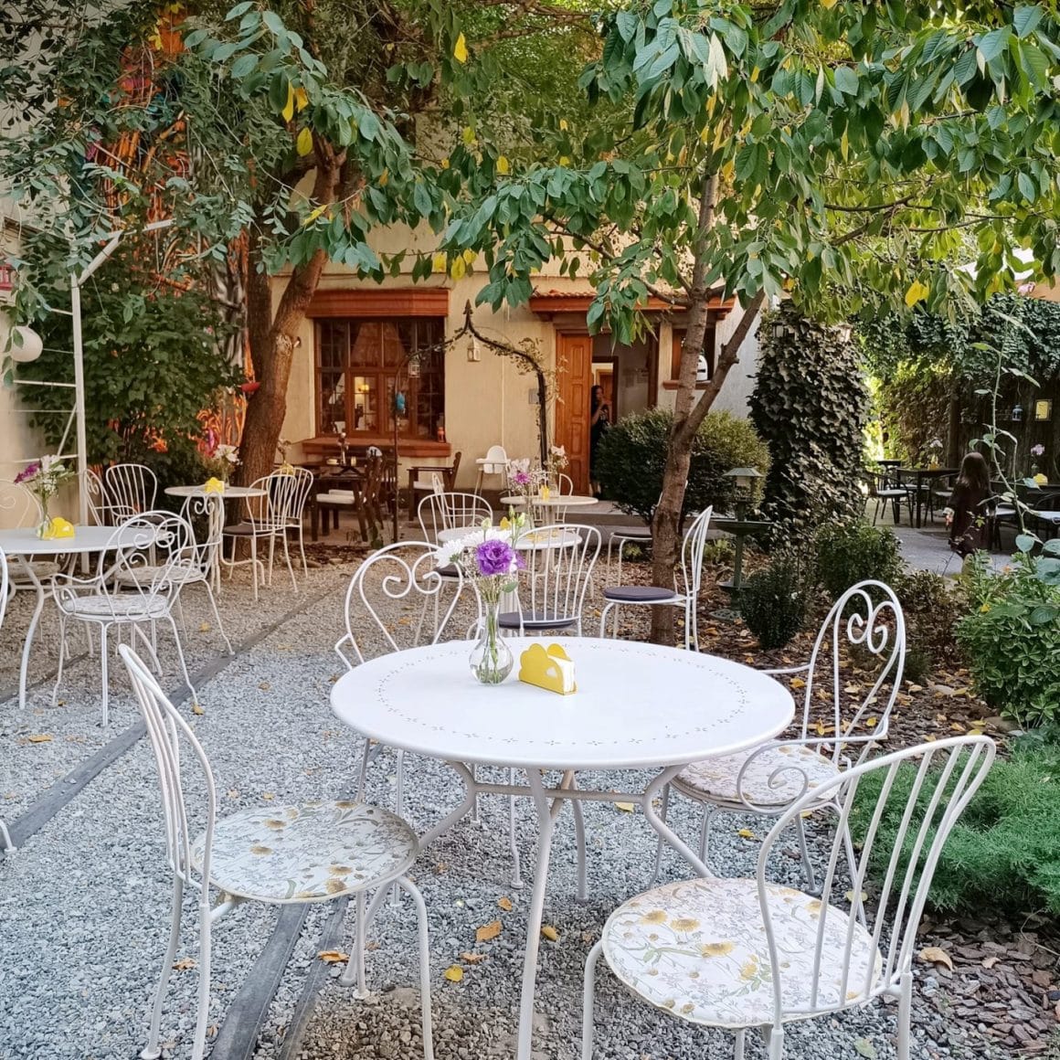 Terasa Greentea din București, cu mese albe, metalice, rotunde, si curtea umbrita de copaci inverziti