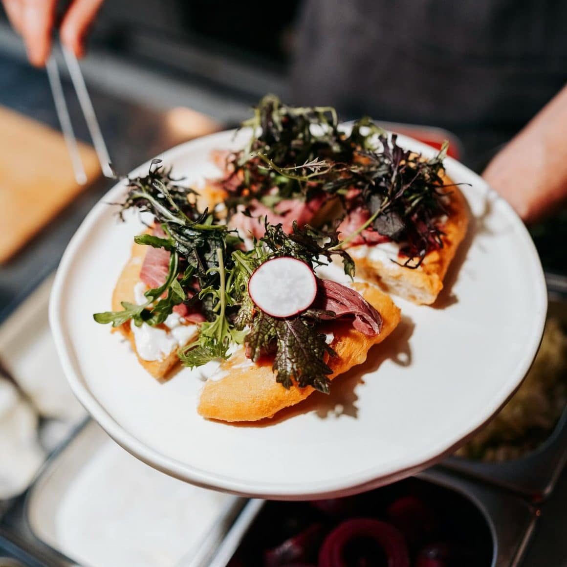 Langoș uns cu cremă de telemea, “decorat” cu felii crud-uscate din ceafă de porc și ridiche marinată de la restaurant Kombinat Sibiu