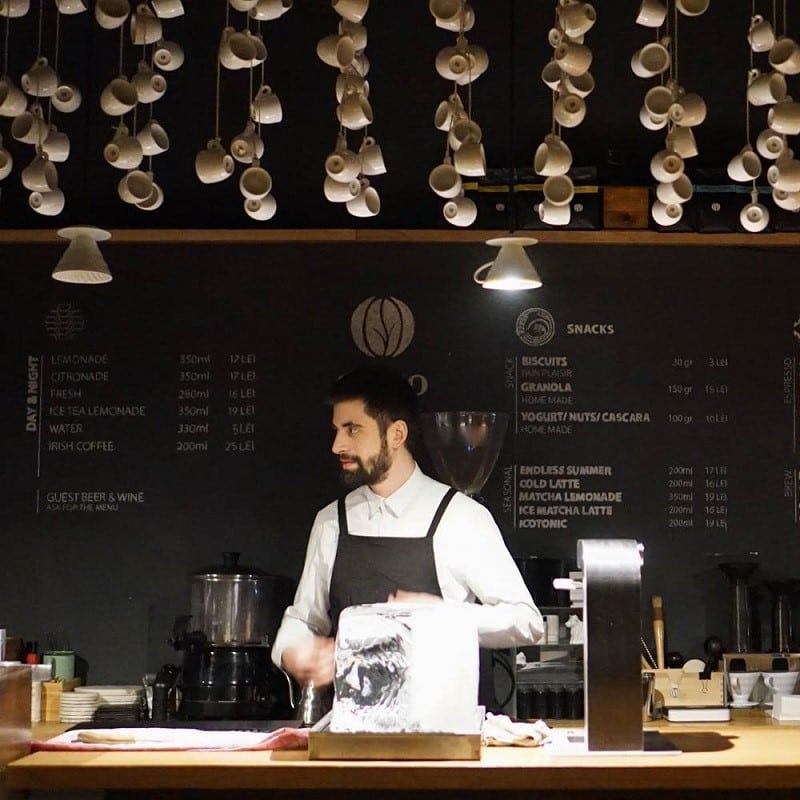 bartista in spatele baruluiu decorat cu cesti de facea de la Origo. cafenele București