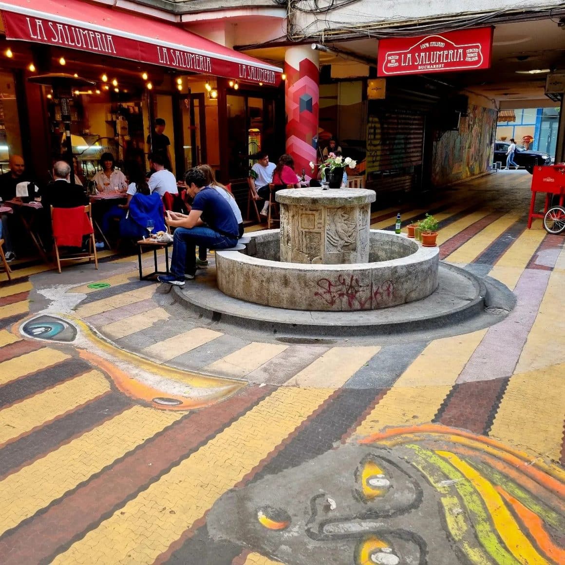 Mai mulți oameni iau masa La Salumeria, pe Claea Victoriei, București, restaurant situat intr-un gand, cu o fantana interioara.