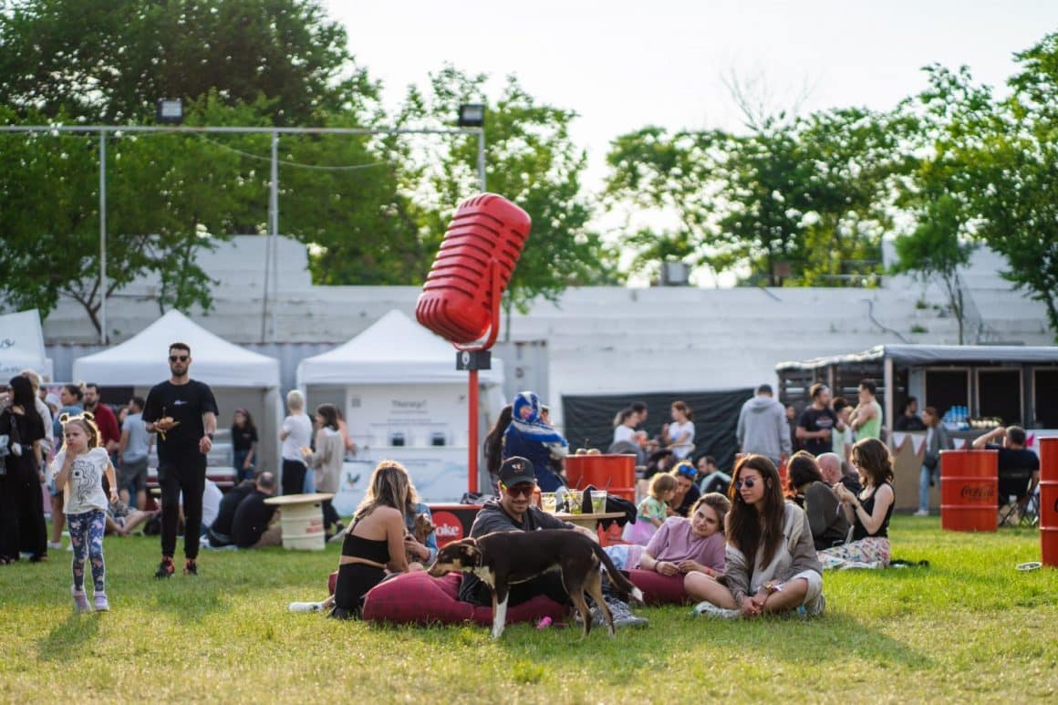 Multi oameni ieșiti la BurgerFest, unii intinsi pe iarba, altii in trecere, se distreaza in aer liber.