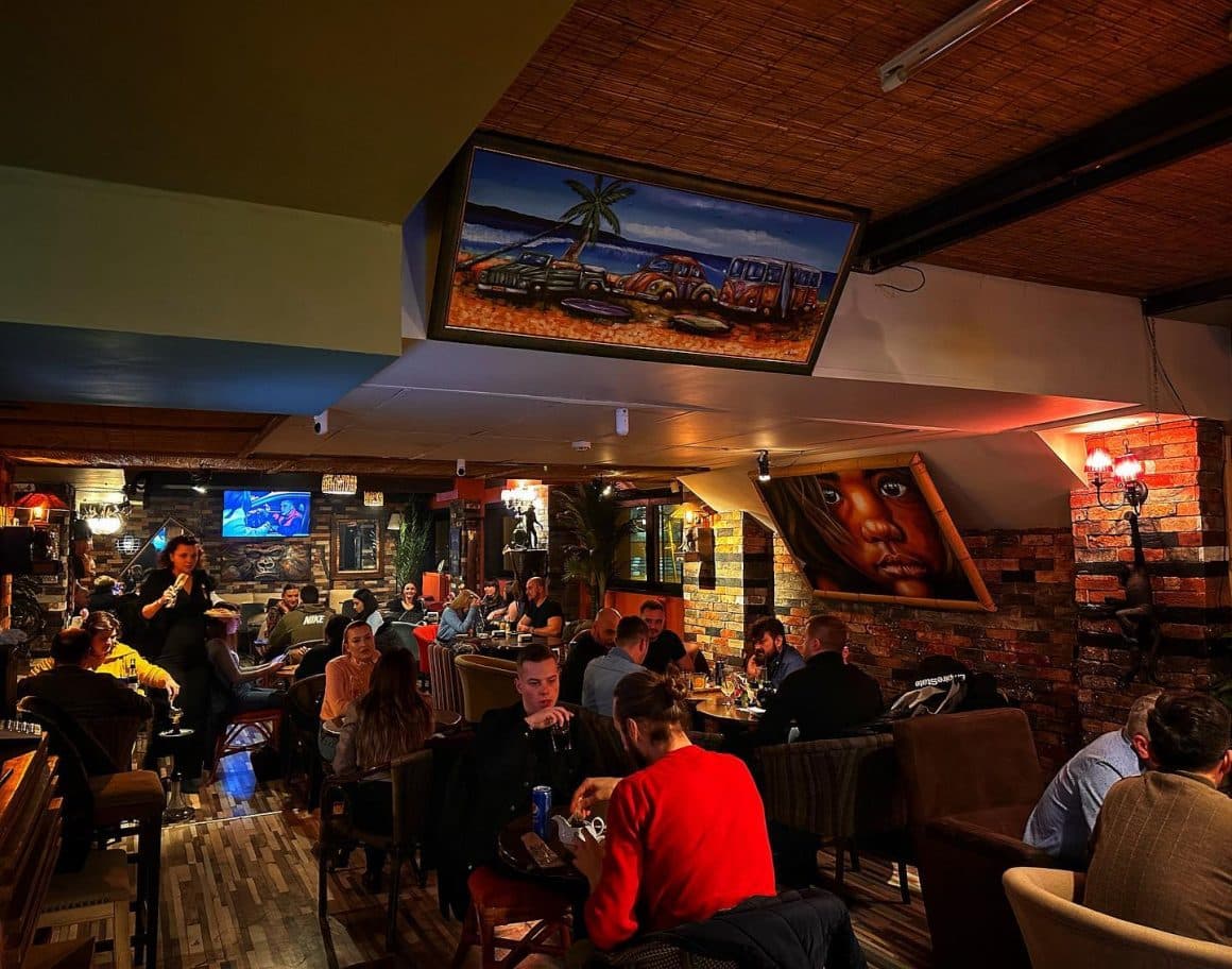 oameni așezați la mese, seara in restaurant Tiki Bistro, cu geamuri mari si draperii colorate, si tavan din lemn iluminat in mai multe culori, unul din restaurante bune din Iași