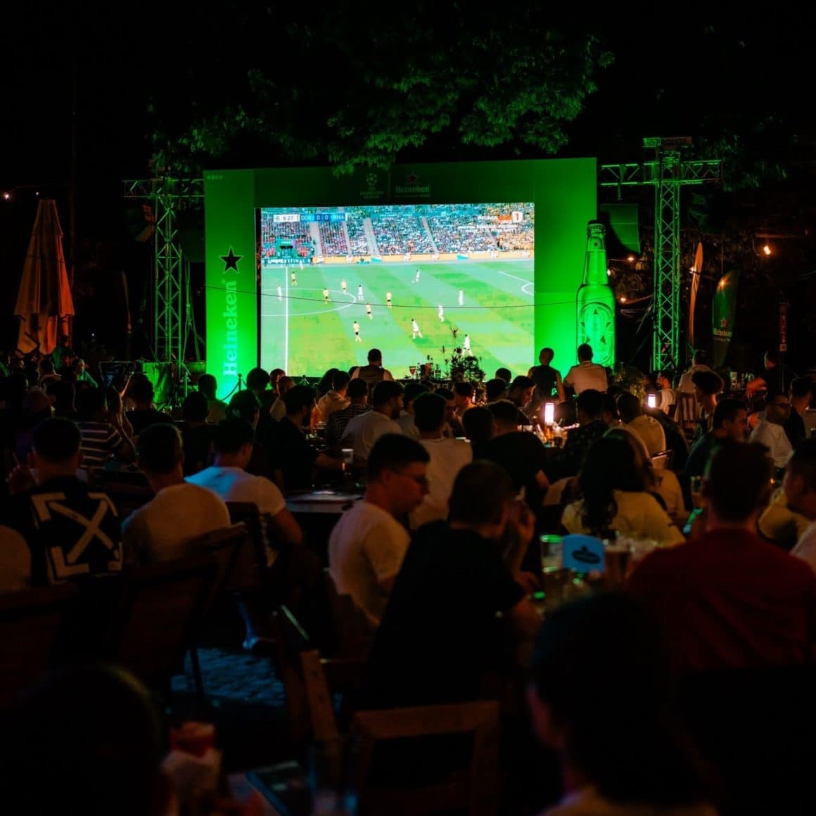 imagine de ansamblu din Beraria H din București, de pe terasă, cu mulți oameni asezati la mese care urmaresc meciuri proiectate pe un ecran gigant.