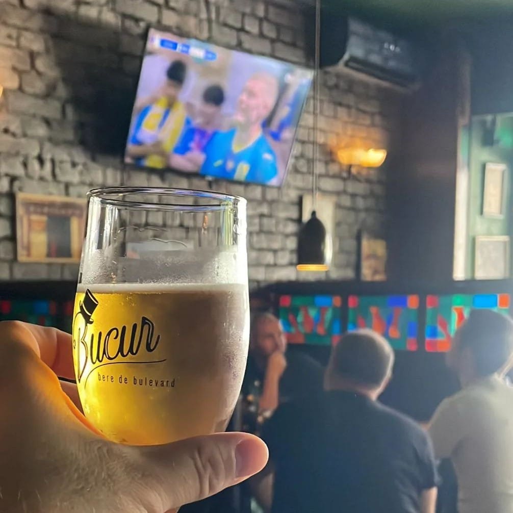 close up cu un pahar d ebere bucur, iar in fundal , blurat, un ecran tv care proiecteaza un meci. terase și baruri sportive București