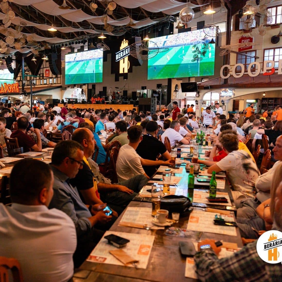 imagine de ansamblu din Beraria H din București, ca o hală imensaă, cu mulți oameni asezati la mese care urmaresc meciuri proiectate pe 2 ecane foarte mari.