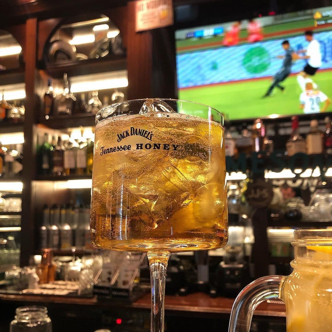 close up cu un pahar de jack daniels, iar in fundal un ecran tv cu meci d efotbal