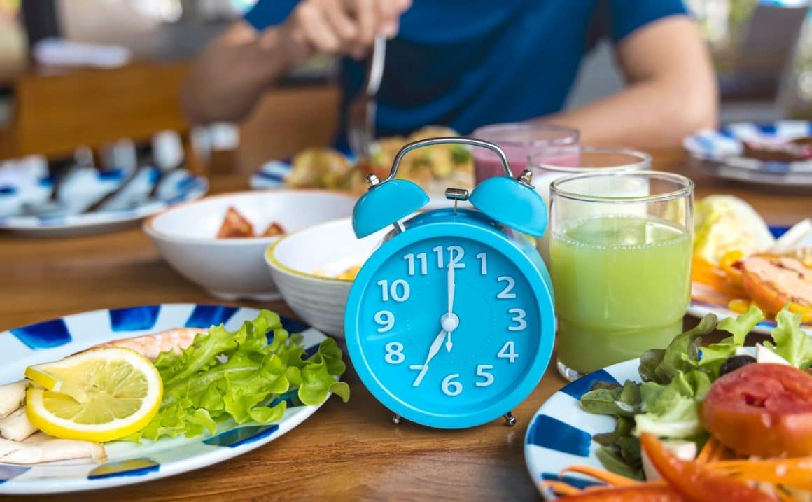Imagine concept pentru post intermitent / fasting: prim plan cu un ceas albastru, alaturi de care sunt farfurii cu mancare sanatoasa, iar in fundal, blurat, un barbat mananca dintr-o farfurie